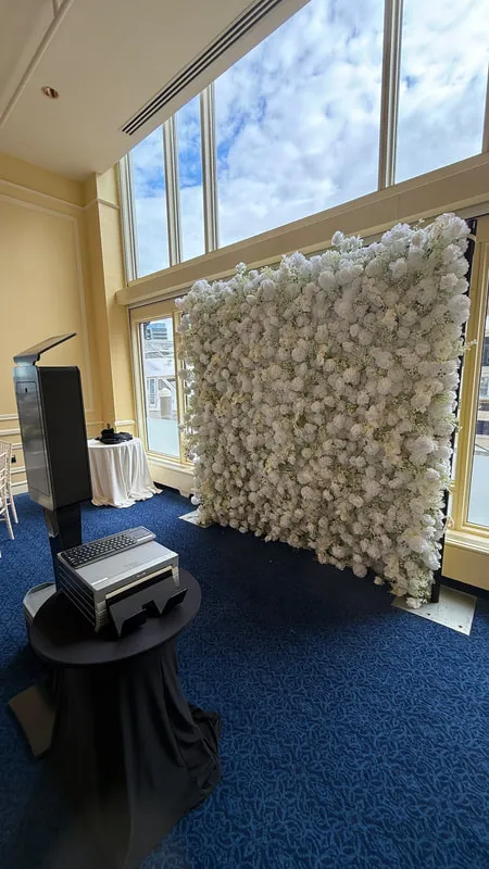 Flower wall photo booth setup beside large venue windows with a white bloom backdrop